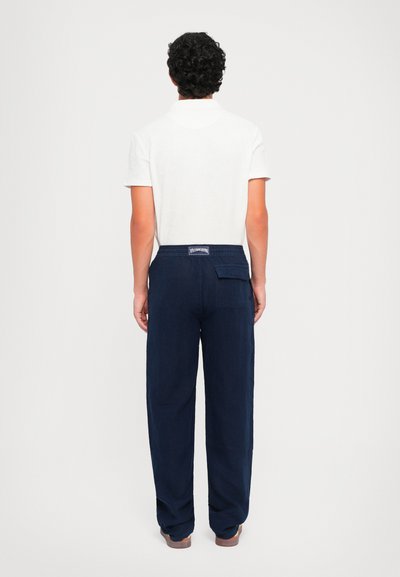 Dunkelblaue Leinenhose mit lockerem Schnitt, elastischem Bund und Gesäßtasche. Getragen mit einem weißen Poloshirt, von hinten betrachtet.