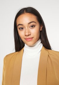 A model wearing a beige blazer over a white turtleneck. The blazer features a smooth texture and structured lapels.