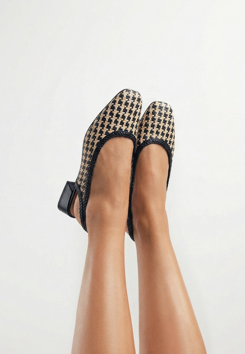 Pies con zapatos slingback de tacón bajo y bloque, con patrón tejido en negro y beige sobre un fondo claro.