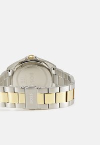 Orologio in acciaio inox con finitura argento e dorata, dotato di bracciale in metallo, che presenta una cassa tonda, una corona testurizzata e un dettaglio del logo sulla fibbia.