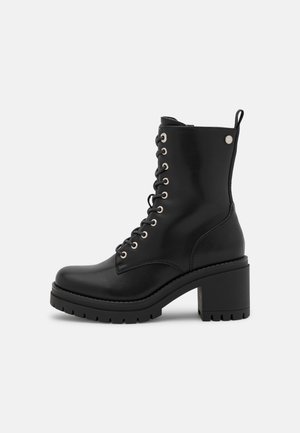 Bottine en cuir noir à talon carré, lacets à l'avant, œillets métalliques et languette à l'arrière sur fond blanc.