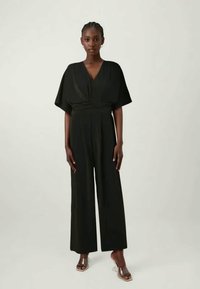 Μαύρο σορτς με φαρδιές γραμμές, βαθύ V-λαιμό και χαλαρά μανίκια, φτιαγμένο από μαλακό ύφασμα. Το μοντέλο φοράει διάφανες σандάλες με τακούνι.