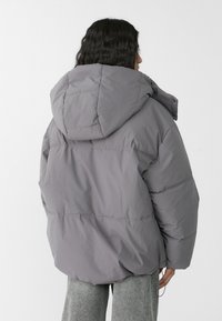 Graue Pufferjacke mit großer Kapuze, gestepptem Design und sichtbaren Nähten. Das Material wirkt weich und leicht glänzend. Sie hat einen lässigen Schnitt.