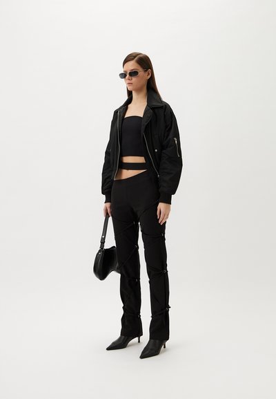 Black bomber jacket layered over a black crop top, paired with fitted black pants featuring a unique lacing design and pointed heels.