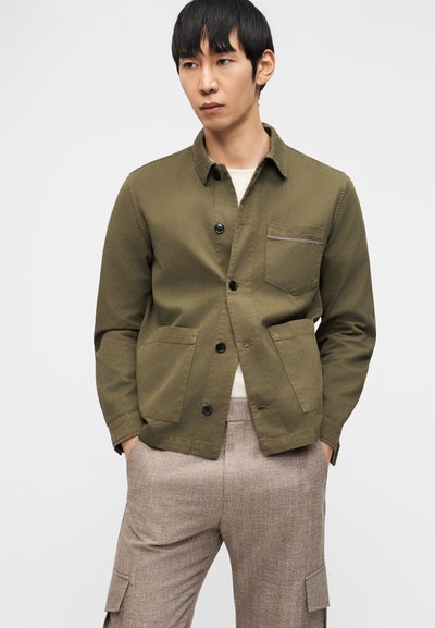 Άνδρας που φοράει χακί κουμπωτό σακάκι με τσέπες και μπεζ παντελόνι cargo, στέκεται με τα χέρια στις τσέπες μπροστά σε λευκό φόντο.