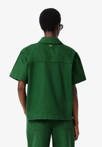 Grünes kurzärmliges Hemd aus strukturiertem Stoff, mit Kragen und einem kleinen Lacoste-Logo nahe dem Ausschnitt auf der Rückseite.