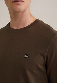 Hnědý bavlněný tričko s kruhovým výstřihem a malou bílou a černou výšivkou vlaštovky na levé hrudi. Hladká textura.