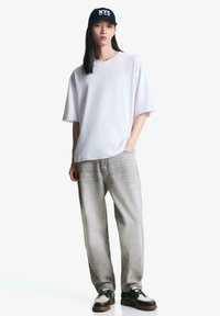 Hvid oversized t-shirt, lysegrå lige ben jeans, sort og hvid sko med brun sål, og en marineblå kasket med "NYC" tekst.