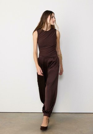 Žena v bezrukávovom tmavohnedom overal s nariasenými nohavicami a zladenými opätkami stojaca pred jednoduchou bielou stenou.