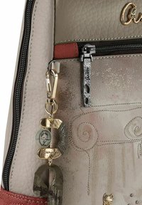 Beige Ledertasche mit roten Akzenten, versehen mit Metallbeschlägen, dekorativen Anhängern und geprägten Details. Reißverschluss für Sicherheit.