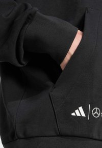 Sweat-shirt noir avec une poche kangourou, arborant des logos blancs d'Adidas et de Mercedes. Fabriqué en tissu texturé avec des poignets côtelés.
