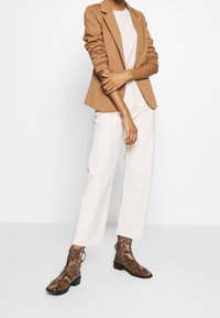 Světle hnědý sleek blazer přes bílý žebrovaný svetr, doplněný volnými krémovými kalhotami a hnědými kotníkovými botami s hadím vzorem.