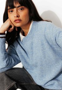 Pull bleu clair en tissu doux et texturé, avec des poignets côtelés et un col rond, superposé sur une chemise blanche à col.