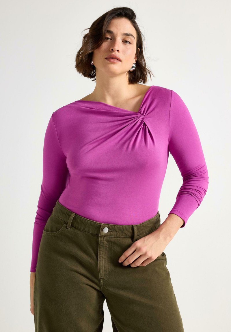 Figursydd långärmad topp i klarrosa med draperad halsringning och veckdetaljer. Kombinerad med olivgröna högmidjade byxor.