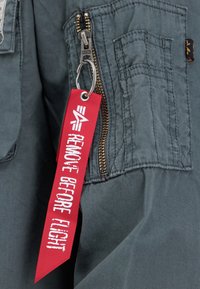 Gray fabric jacket with a gold zipper and a red "REMOVE BEFORE FLIGHT" tag attached to a keychain, featuring additional stitching details.