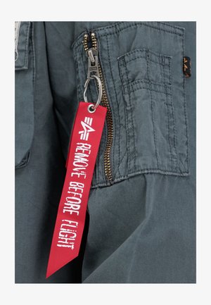 Veste en tissu gris avec une fermeture éclair dorée et une étiquette rouge "ENLEVER AVANT LE VOL" attachée à un porte-clés, avec des détails de couture supplémentaires.