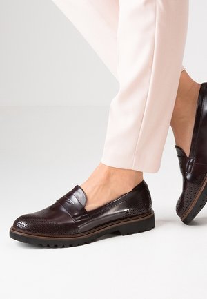 Donkerbruine leren brogue loafers gedragen met lichtbeige broek, met gedetailleerde perforaties en dikke zwarte rubberen zolen.