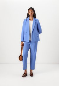 Light blue tailored suit jacket with two buttons, matching trousers, and brown loafers. Beige handbag with a shoulder strap. Textured white top.
