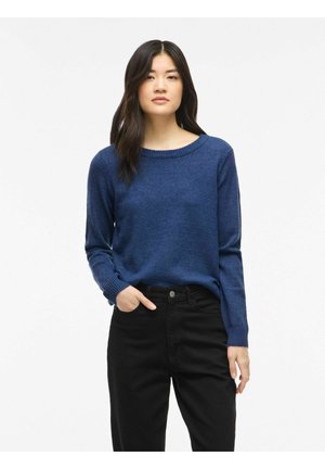 Femme aux longs cheveux foncés portant un pull bleu et un pantalon noir, debout avec une main dans sa poche devant un fond blanc uni.