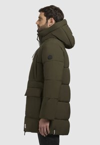 Olivengrøn pufferjakke med hætte, der har en lynlåslomme, sidelommer og et logo-patch på ærmet. Tekstureret, quiltet design.