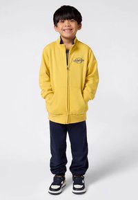 Gelber Sweatshirt mit Reißverschluss und Seitentaschen, mit einem Logo auf der Brust. Kombiniert mit marineblauen Jogginghosen und weißen Turnschuhen mit schwarzen Akzenten.