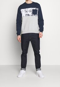 Modelo masculino alto vestido con un sudadera en tonos azul marino y gris con un patrón floral, combinado con jeans de mezclilla oscuros y zapatillas deportivas blancas, de pie contra un fondo liso.
