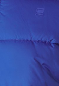 Spilgti zila pūkainā jakas audums ar iešūtu struktūru. Uz virsmas ir viegli reljefā, nedaudz tumšākā zilā krāsā iegravēts "RAW" logotips.