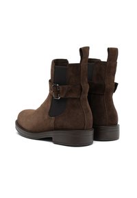 Botas de tobillo de ante marrón con correas de hebilla en los laterales, paneles elásticos negros, tiradores en la parte posterior y tacones bajos en forma apilada sobre un fondo blanco.