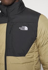 Veste matelassée avec une partie supérieure noire et une partie inférieure kaki. Fermeture éclair, col montant, et logo blanc sur la poitrine.