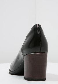 Talons en cuir noir avec un bout arrondi, présentant une texture lisse et un talon bloc en bois stratifié. Coutures visibles le long des côtés.