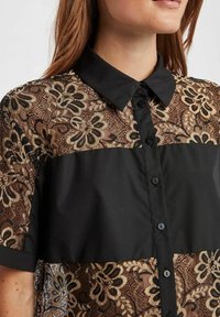 Zwarte blouse met knopen en een bloemenkantoverlay in bruine tinten. Heeft een kraag en korte mouwen, waardoor gestructureerde en transparante texturen worden gecombineerd.