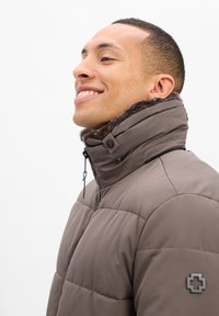 Ein lächelnder Mann trägt eine hochkragen Taupe Daunenjacke mit einer mit Fell gefütterten Kapuze und einem Logo-Patch am Ärmel, blickt nach links.