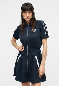 Αθλητικό φόρεμα σε ναυτικό μπλε με φερμουάρ, κοντά μανίκια, ελαστική μέση, λευκές λεπτομέρειες στις τσέπες και λογότυπο στο στήθος. Μαλακό ύφασμα.