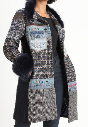 Femme portant un manteau long à motifs avec des poches en denim, une broderie colorée et un bord en fourrure foncée sur le col et les poignets.