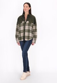 Camisa de cuadros en verde y beige con botonadura frontal, dos bolsillos en el pecho y mangas largas; combinada con jeans de mezclilla oscuros y zapatillas beige.