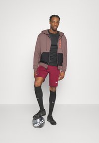 Sports outfit med en bordeaux og sort zip-up hoodie, sort t-shirt, bordeaux shorts, knælange sorte sokker og sorte sportssko.