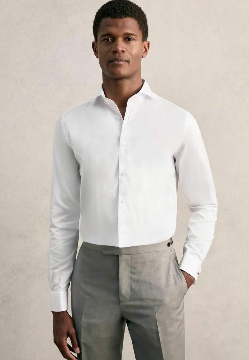 Valge pikkade varrukatega pluus, millel on nööbitav krae, sile tekstuur, kombineeritud heledate hallide kohandatud pükstega, millel on küljetaskud ja nööbid.