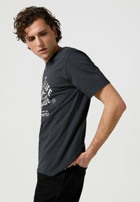 Camiseta de algodón de manga corta en gris oscuro, con un estampado gráfico blanco en la parte delantera y un diseño de cuello redondo. Texturizada con un acabado suave.