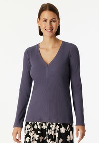 Woman wearing a long-sleeve purple V-neck top with three buttons and a black-and-white floral skirt, smiling against a white background.