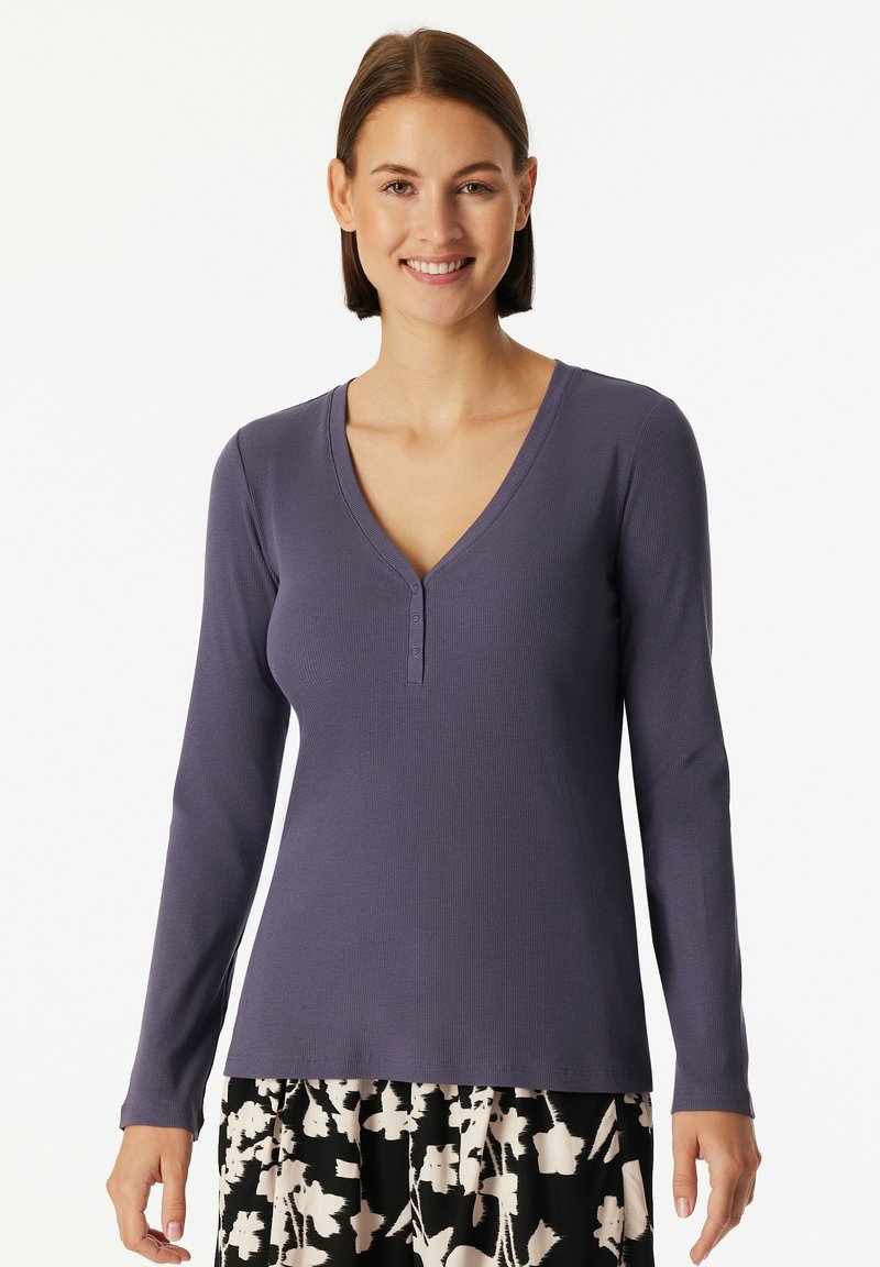 Woman wearing a long-sleeve purple V-neck top with three buttons and a black-and-white floral skirt, smiling against a white background.