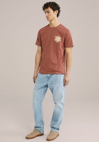 Roestkleurig t-shirt met een bedrukt logo op de borst, gecombineerd met lichtblauwe, losse jeans en beige sneakers.