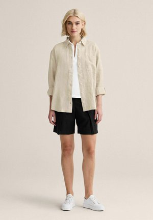Lyshåret kvinde iført beige skjorte med knapper over hvid top, sorte shorts og hvide sneakers, stående foran en ensfarvet beige baggrund.