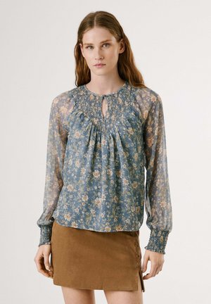 Bluse mit blauem Blumenmuster, langen Ärmeln, gerafftem Kragen und Schlüssellochöffnung. Kombiniert mit einem braunen Wildlederrock mit seitlichen Schnürungen.