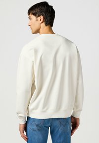 Sudadera color crema con un cuello redondo, puños y dobladillo acanalados, hecha de una tela suave, combinada con jeans azules con detalles de costura angulares.