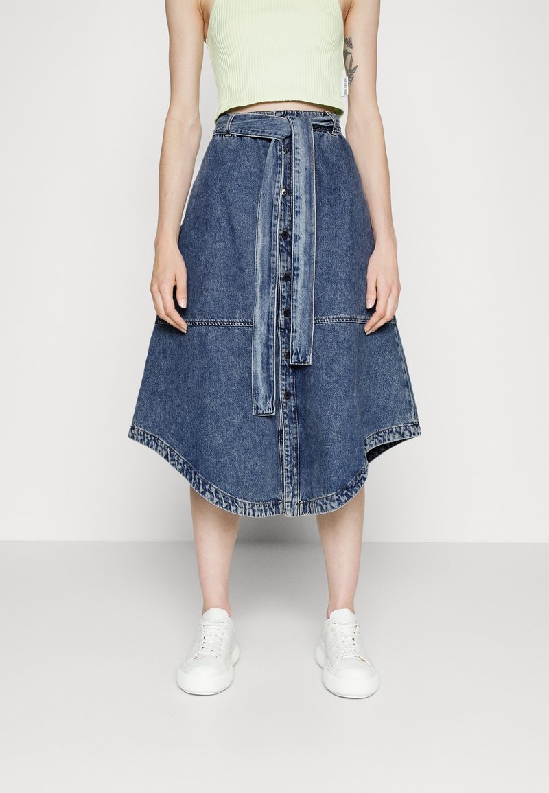 Denim midi sukně s rozšířeným lemem, s páskem v pase, předními knoflíky a dvoubarevným designem. V kombinaci s bílými teniskami.