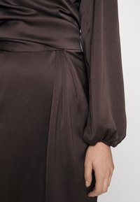 Vestido marrón texturizado con un diseño drapeado, mangas largas abullonadas y detalles fruncidos en la cintura. Tejido suave con un brillo sutil.