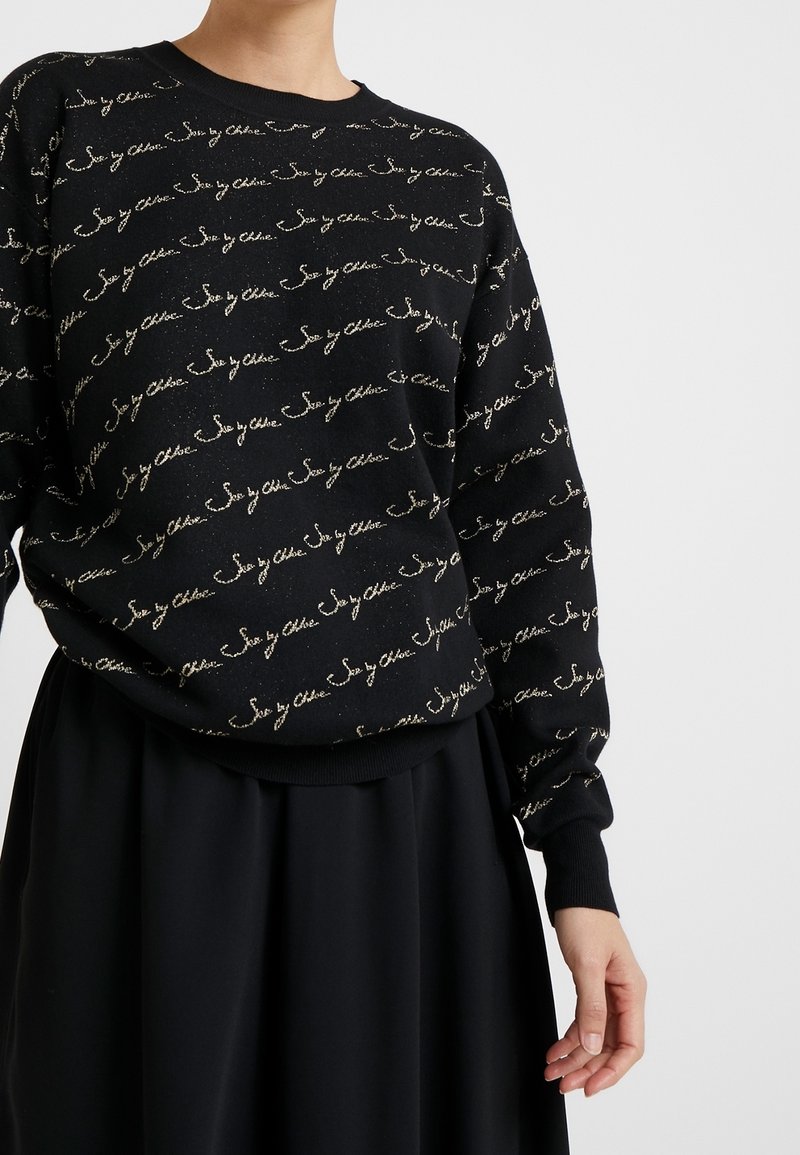 Czarny sweatshirt z powtarzającym się beżowym napisem "See by Chloe", noszony z czarną spódnicą, częściowy widok osoby od szyi do ręki.