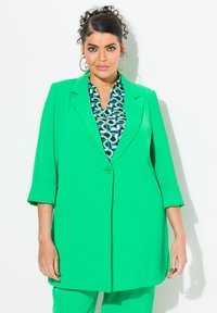 Zelený blazer s jedním knoflíkem a ohrnutými rukávy, v kombinaci s vzorovanou blůzou se vzory kruhů v modrých a zelených odstínech. Hladká textura.