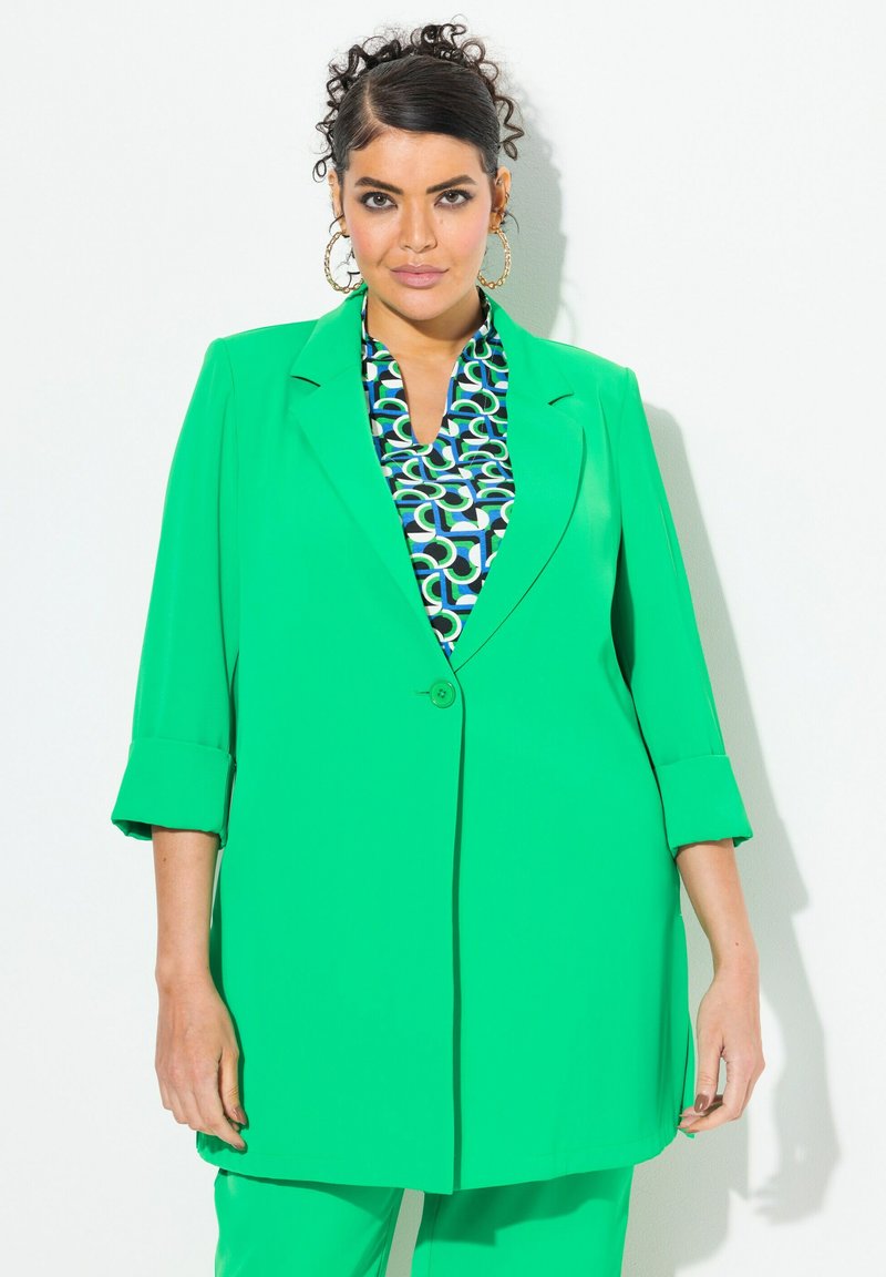 Zelený blazer s jedním knoflíkem a ohrnutými rukávy, v kombinaci s vzorovanou blůzou se vzory kruhů v modrých a zelených odstínech. Hladká textura.