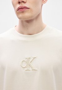 Μπεζ βαμβακερό t-shirt με υφασμάτινο κρεμ κεντημένο λογότυπο "cK" στην αριστερή πλευρά. Στρογγυλή λαιμόκοψη και κοντά μανίκ�ια. Λείος ύφασμα.